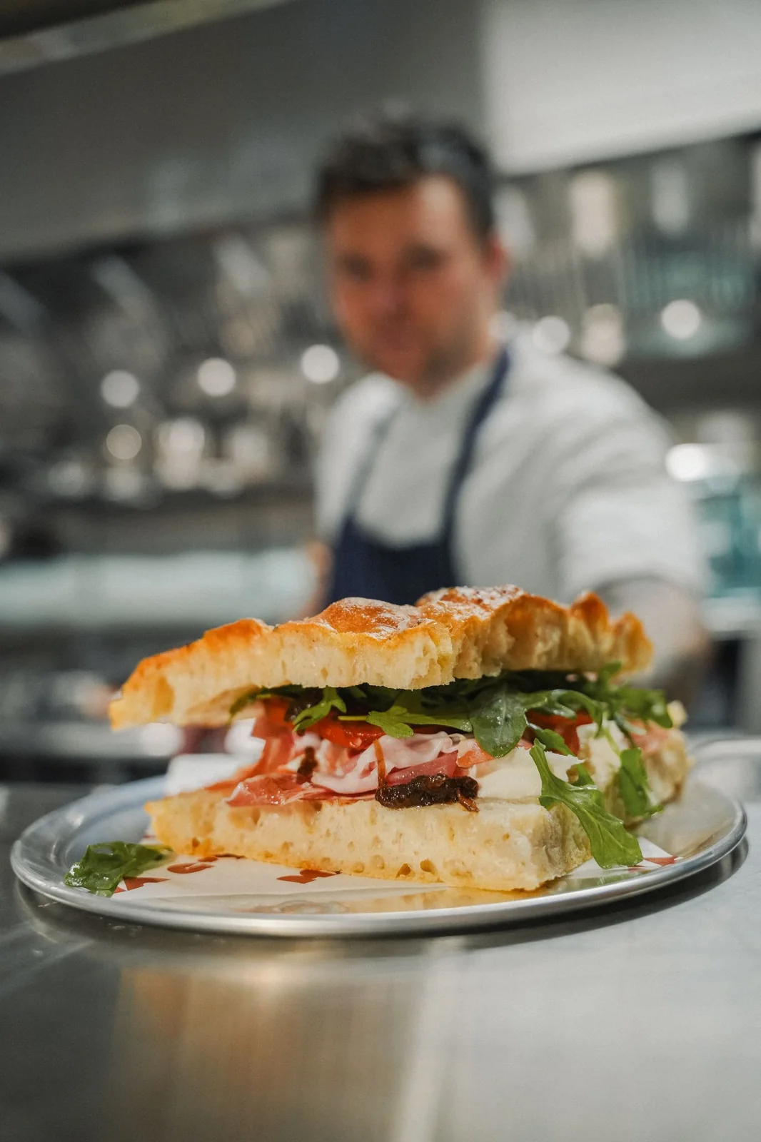 A chef presenting a plated focaccia sandwich on a tray at the Mamma G's pass