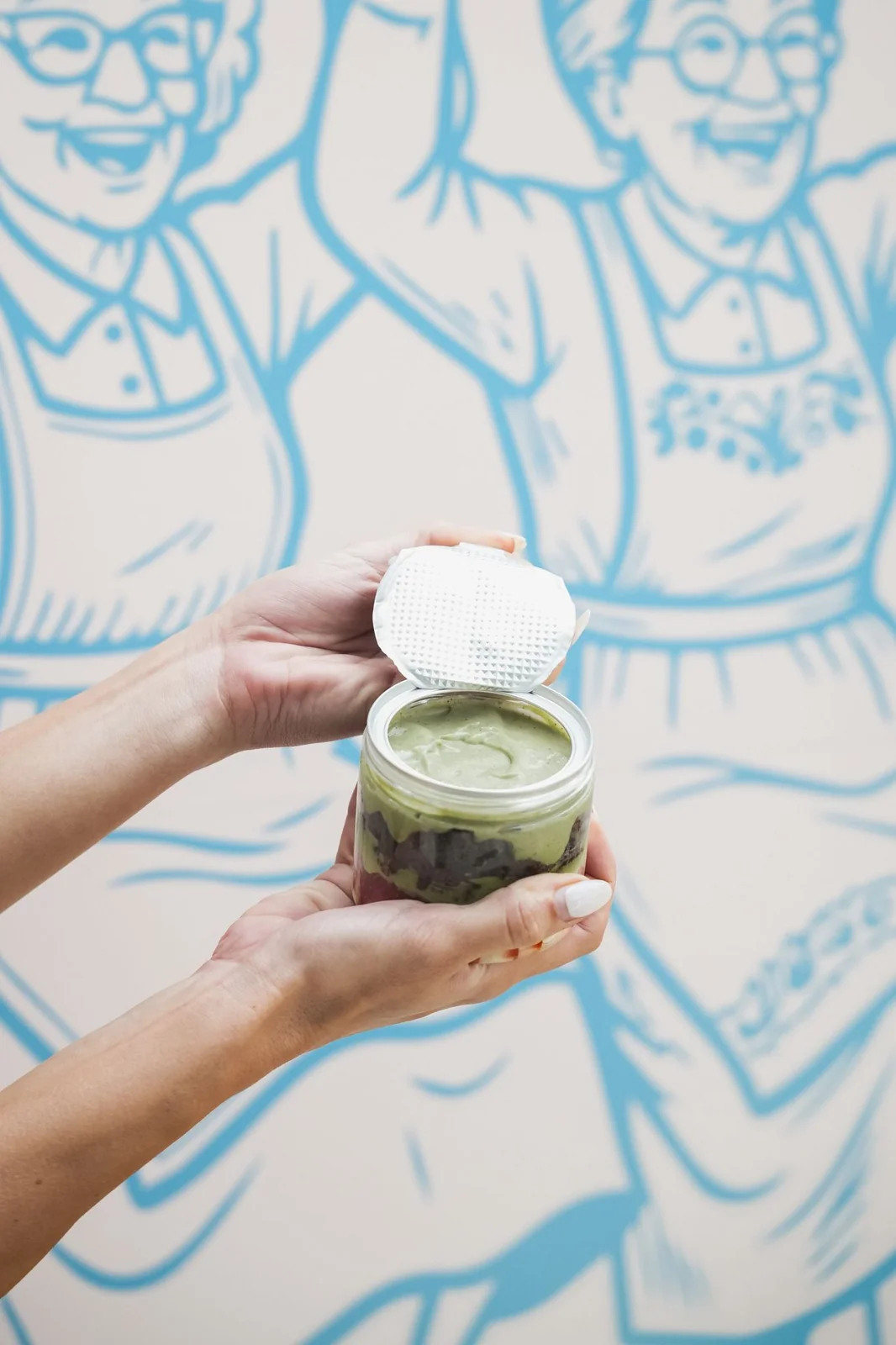 A hand passing over a small jar of matcha dessert in front of the blue nonna mural